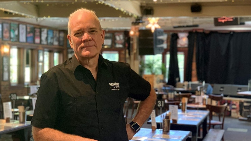 A man standing at a bar table with his hand on his hip, in front of rows of empty tables inside a bar.