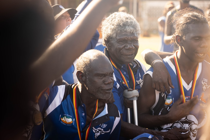 Three men, two old and one young, are wearing blue sports shirts with medals around their necks. They are happy.