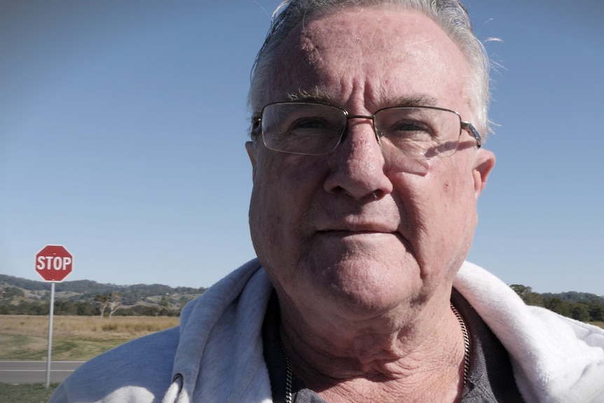 A head shot of an older man with glasses looking past the camera with a stop sign in the background