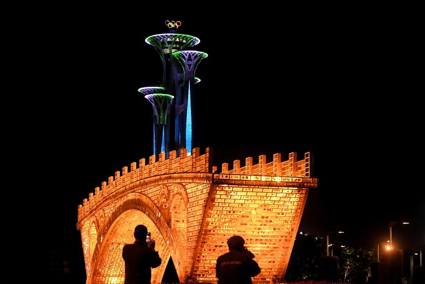 Two people silhouetted against an illuminated bridge installation in Beijing at night take pictures.