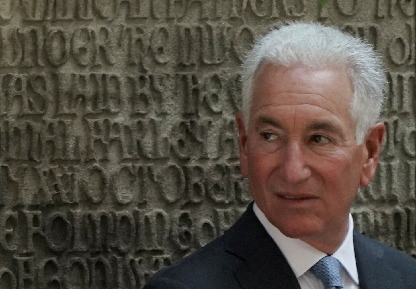 A closeup of an older white man with white hair, smirking slightly next to a carved wall