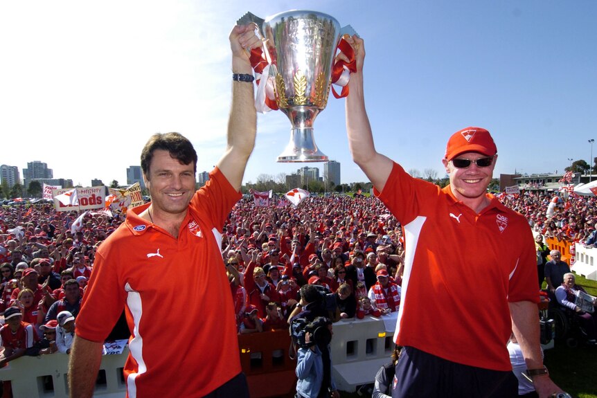 Two guys in red clothing holding up a premiership cup with the sun shining in the background