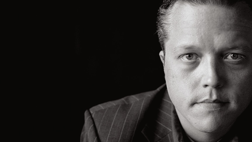 Black and white photo of Jason Isbell wearing a pinstripe jacket against a black background