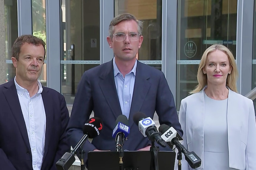 an older man standing next to a younger man wearing glasses standing next to a woman all talking to the media