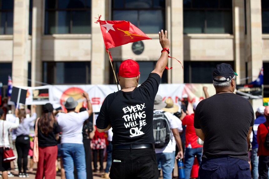 A man holds a flag in the air as he is pictured from behind wearing a shirt stating 'everything will be ok'