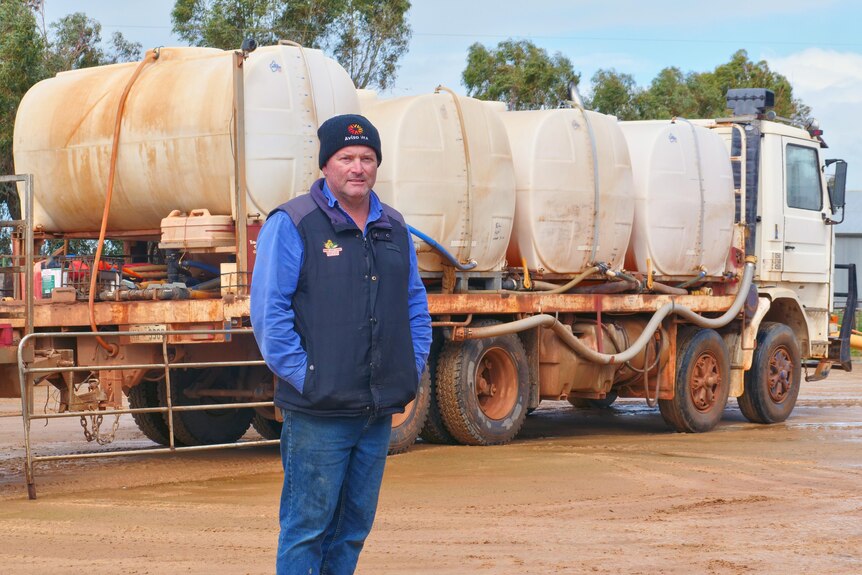 A man in blue jeans, long sleeve shirt, puffer vest and beanie stands in front of a truck carrying chemical tanks.