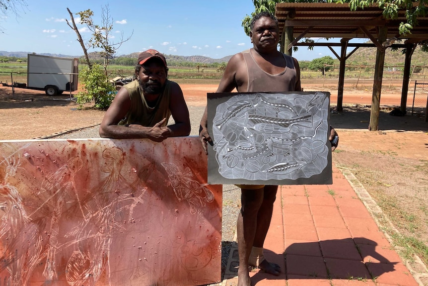 Shaun Namarnyilk and Gabriel Maralngurra with recently finished artworks. They are outside, and there is red dirt and trees.