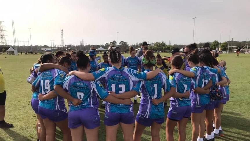 A group of girls huddle in their blue and purple jerseys with their backs to the camera on an oval