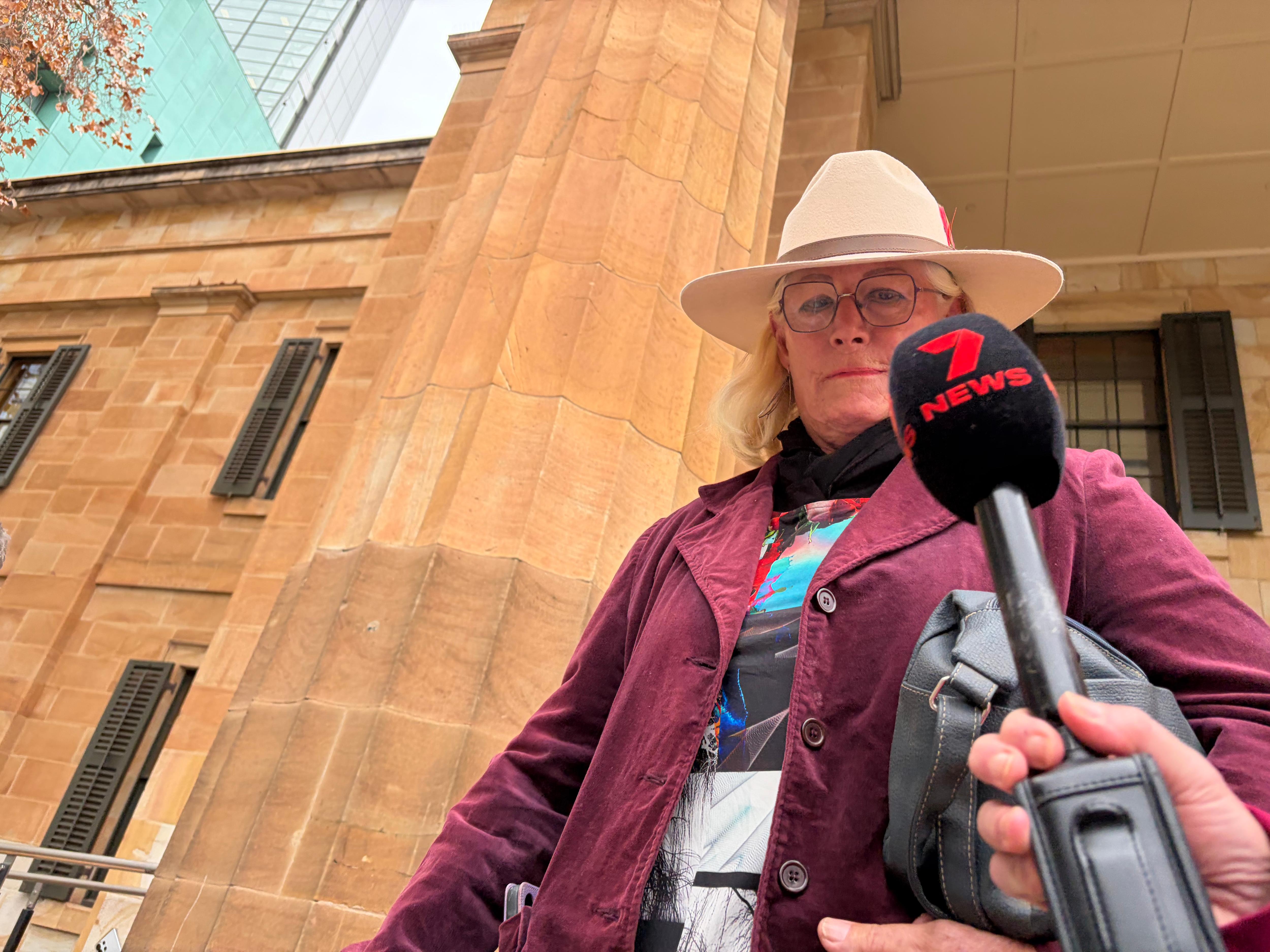 A person wearing an Akubra hat and maroon jacket with a 7 news microphone near their face in front of a stone building