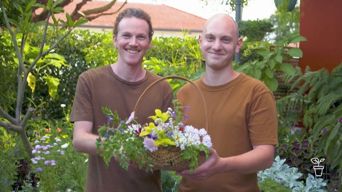 two men in a garden smiling at the camera and holding a basket filled with cut flowers
