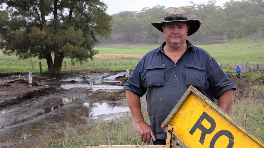 Man in workshirt and hat stands next to a muddy roadway