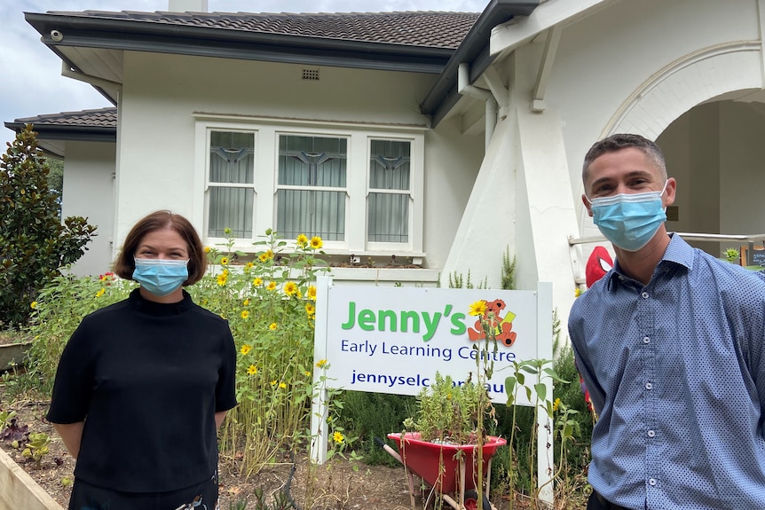 A woman and man wearing face masks stand a metre away with a 'Early Learning Centre' sign in the middle