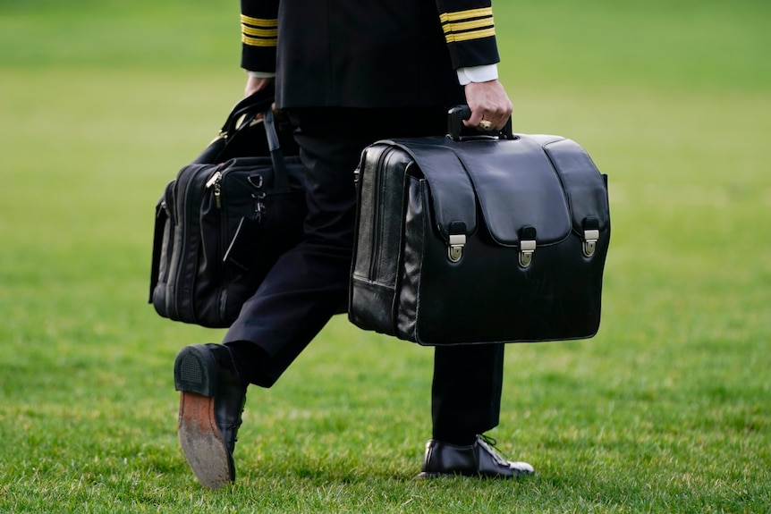 A waist-down photo of a person who is wearing a uniform carrying a big leather bag in each hand