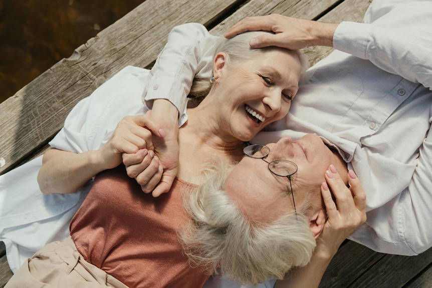 elderly man and woman are lying on a pier hugging holding each others facies smiling they both have grey hair 