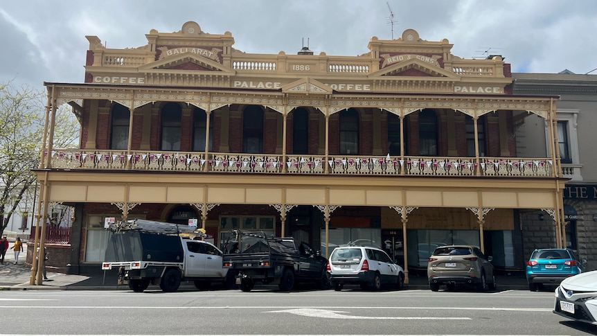 A 1800's cream and red building with latticework across the verandah, cars parked in front.