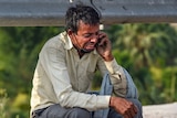 A man with a face mask under his chin sobs while sitting on the roadside with a phone to his ear
