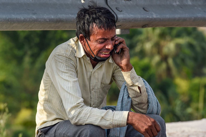 A man with a face mask under his chin sobs while sitting on the roadside with a phone to his ear