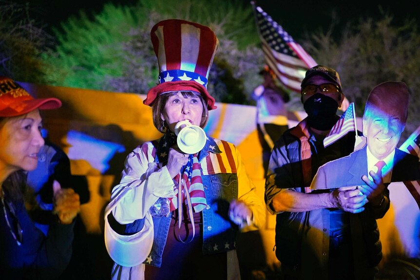 A woman in a US flag outfit and Uncle Sam hat speaks into a megaphone in front of a protest