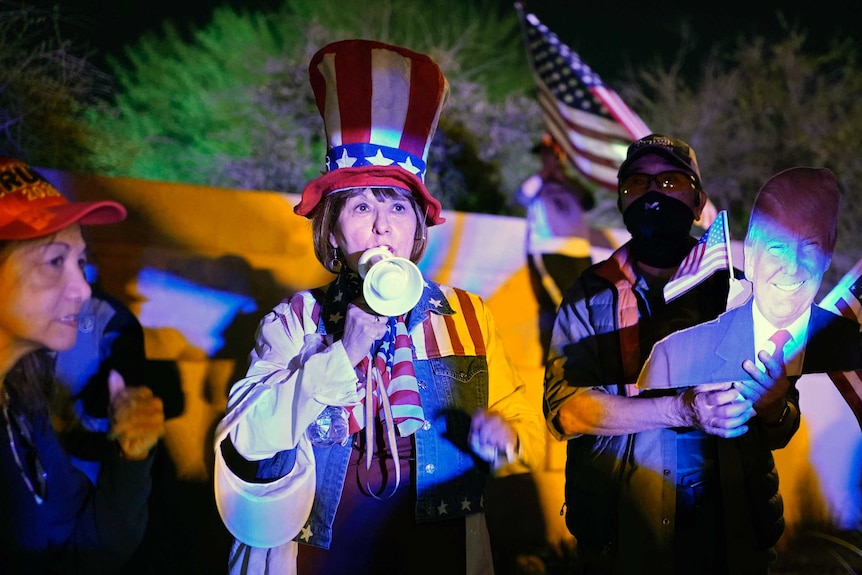 A woman in a US flag outfit and Uncle Sam hat speaks into a megaphone in front of a protest