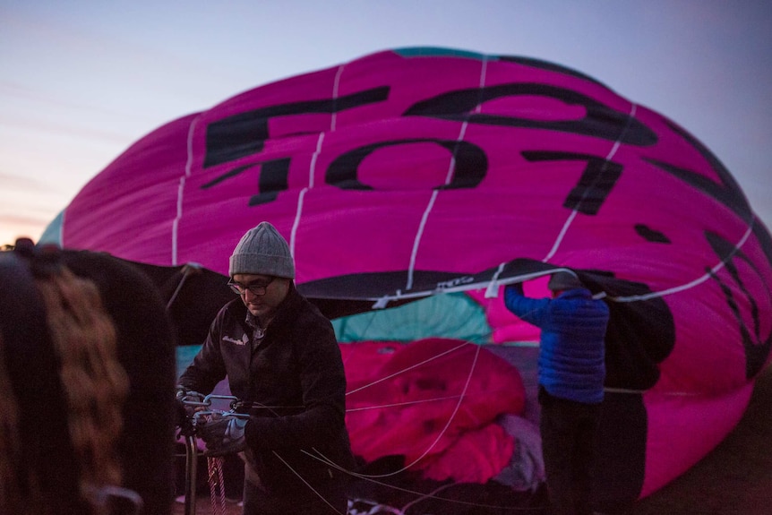 In dawn light, a hot air balloon lies on its side, inflating, as Chris attends to rigging on the basket.