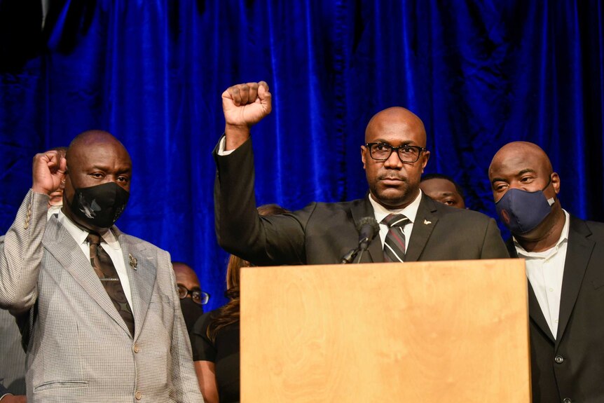 Two black men in suits and ties raise their right arms in triumph in front of blue curtain in room.