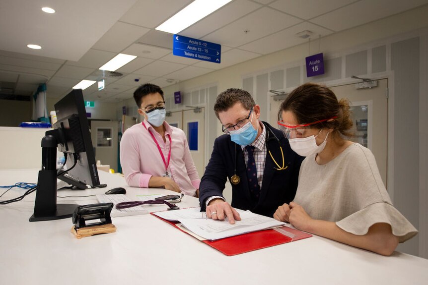 Man wearing a suit stands between two junior doctors, looking at paper work.
