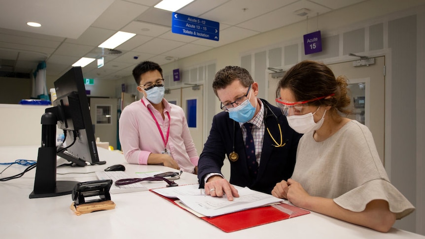 Man wearing a suit stands between two junior doctors, looking at paper work.