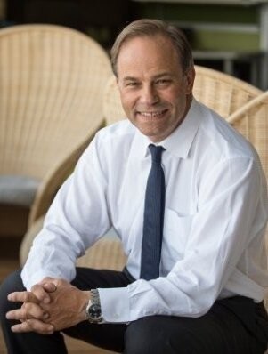 A middle-aged man in business attire sits in a chair, smiling.