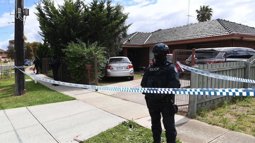 A police officer wearing body armour, helmet and face mask stands behind police tape outside a house