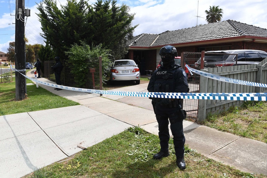 A police officer wearing body armour, helmet and face mask stands behind police tape outside a house