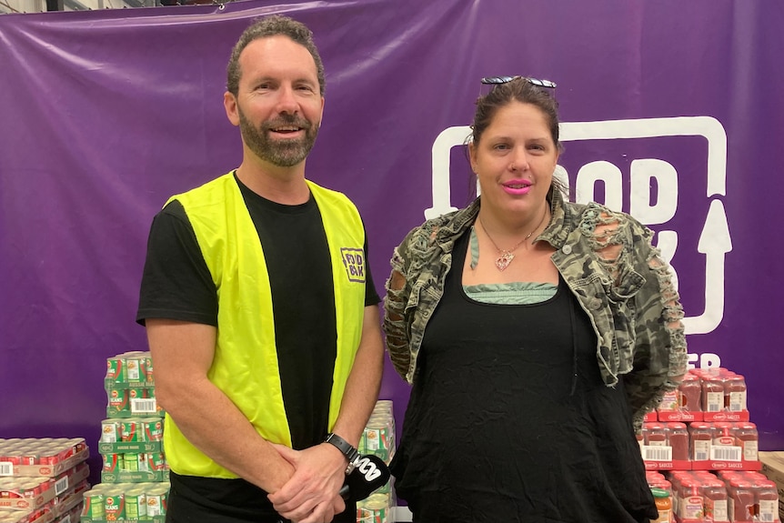 A man and a woman stand in front of stacks of canned food. The man wears a high-vis vest, the woman has bright pink lipstick 