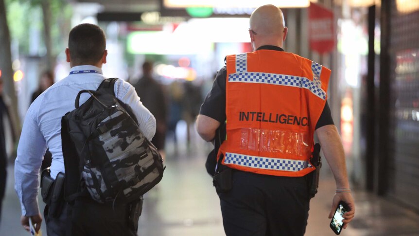 View of the back of a man carrying a backpack (left) and a man (right) wearing an orange vest with 'Intelligence' written on it