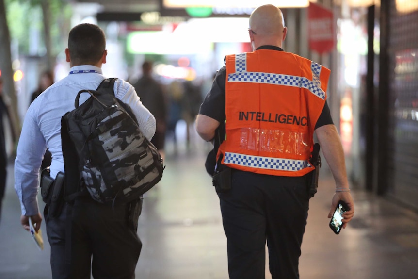 View of the back of a man carrying a backpack (left) and a man (right) wearing an orange vest with 'Intelligence' written on it