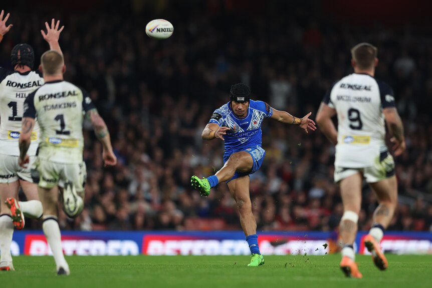 A Samoan rugby league player eyes the ball as he extends his left foot after kicking for goal while England players watch. 