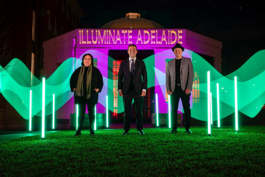 Three people stand in front of an illuminated hut with the words Illuminate Adelaide above it