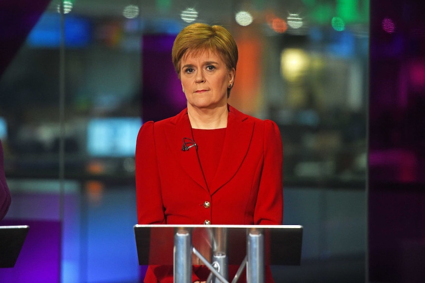 Nicola Sturgeon stands behind a metal lectern in a red pant-suit during a leaders debate.