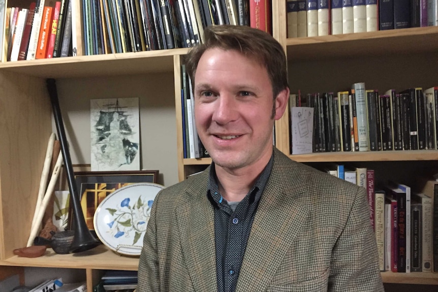 A man in a checked jacket and spotted shirt sits in front of a bookcase.