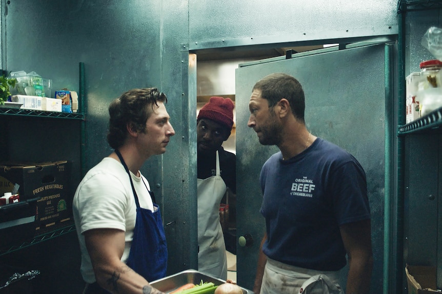 Two young white men stare each other down in a commercial refrigerator, a Black man stands in the doorway behind them