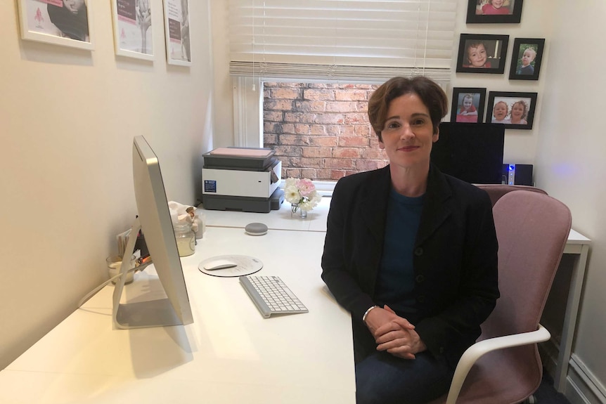 Nicole Highet sits at a desk with an Mac computer in a predominantly white office with photo frames of children on the wall.