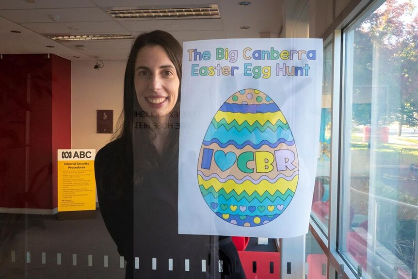Elise Fantin smiles from inside the ABC studio, holding a sign up with a coloured egg on it that says "Easter Egg Hunt".