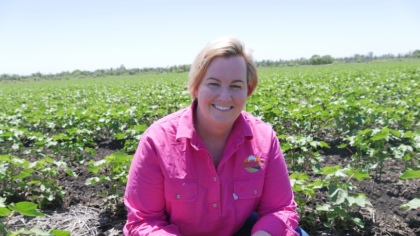A blonde woman in a pink work shirt kneels in a newly planted cotton crop. 