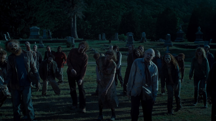 A large group of zombies wearing human clothing stagger through graveyard at night time in a graveyard.