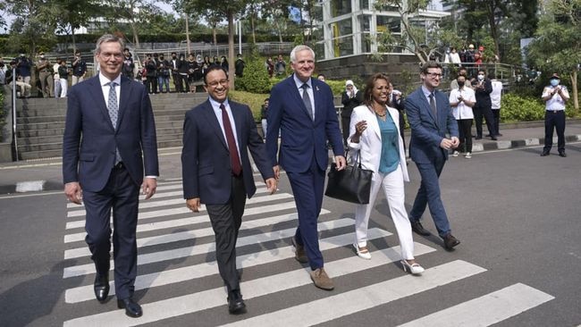 Four men in blue suits and a woman in a white blazer walk parallel to the zebra crossing smiling at the camera.