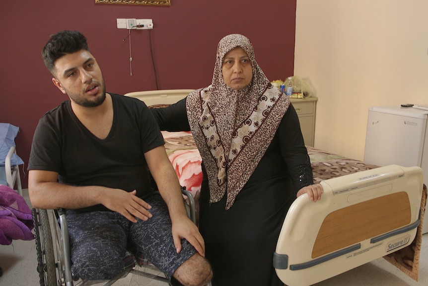 Ghadban sitting in a wheelchair next hospital beds. A woman sits next to him.