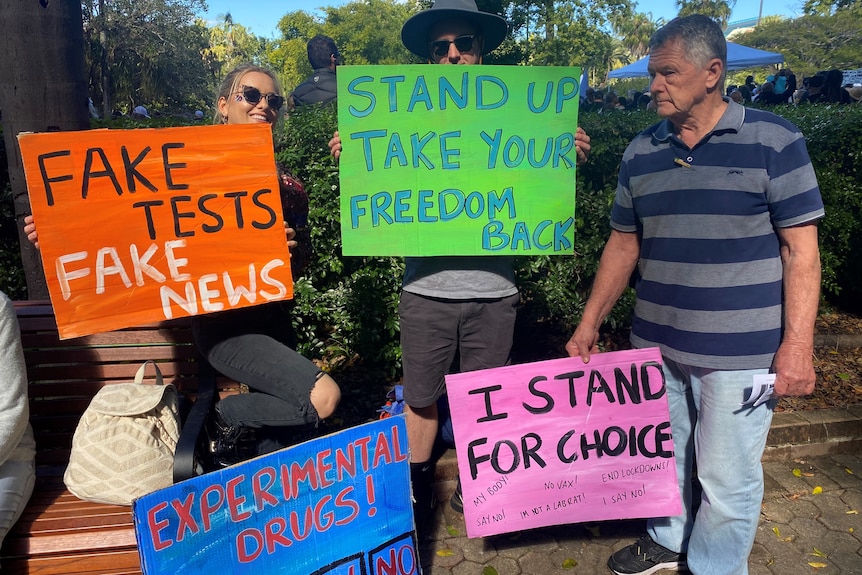 Protesters hold signs at a rally saying 'I stand for choice', 'fake tests fake news', 'stand up take your freedom back'