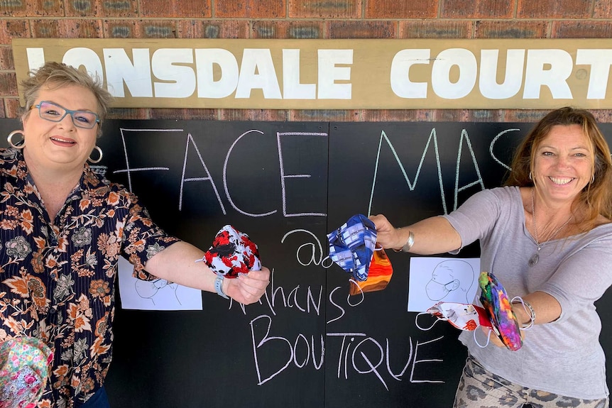 Two women stand holding face masks outside a shop.