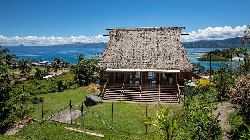 You view a modern take on traditional Fijian bure architecture sitting on a green hilltop overlooking glittering blue waters.