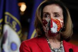 Nancy Pelosi, wearing a red blazer over a grey top with pearls and a patterned face mask.