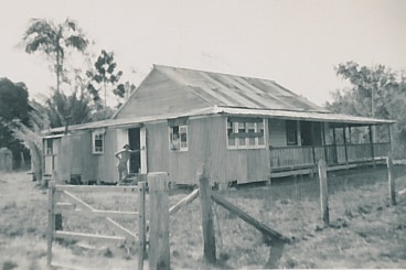 A black and white photos of the wooden pioneer building. A woman stands at the door and looks at the camera.