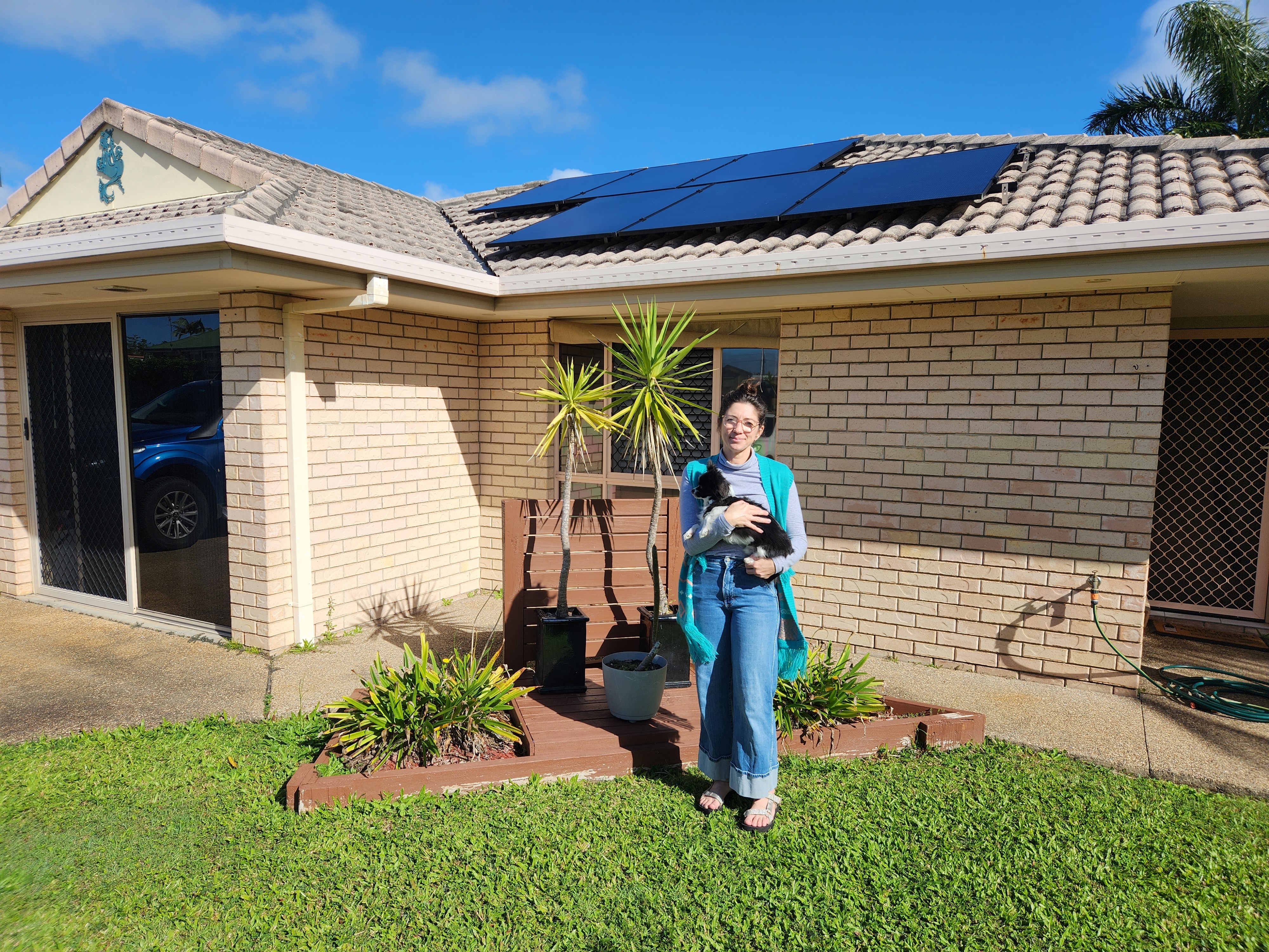 A woman holding a dog, standing in front of a garden at the front of her home.